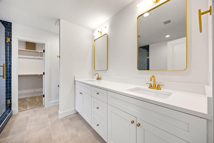 Luxurious primary bathroom featuring a double-sink vanity with quartz countertops, white cabinetry, and elegant gold fixtures. Large framed mirrors, recessed lighting, and a clean modern design create a spa-like atmosphere with direct access to the walk-in closet.