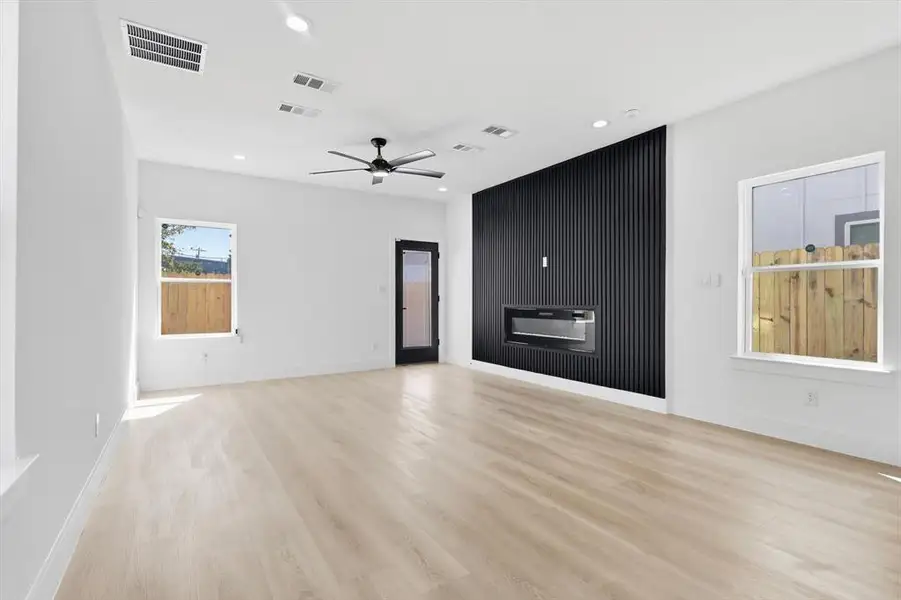 Spacious interior featuring wide plank wood-finish flooring, a modern black accent wall with an integrated linear fireplace, and a ceiling fan with integrated lighting Spacious interior featuring wide plank wood-finish flooring, a modern black accent wall with an integrated linear fireplace, and a ceiling fan with integrated lighting