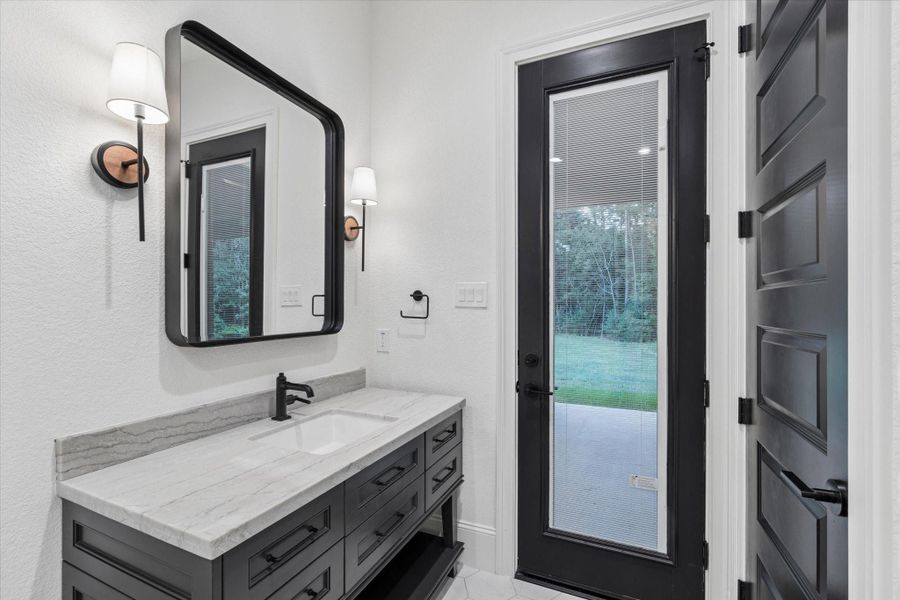 This beautifully appointed pool bathroom continues the home’s sophisticated design with a custom vanity, stone countertop, oversized framed mirror, and coordinated black fixtures. A glass-paneled exterior door provides direct access to the covered patio and backyard, allowing guests to enter without moving through the main living areas. Designer sconces, elevated millwork, and timeless finishes create a polished and functional space perfectly.