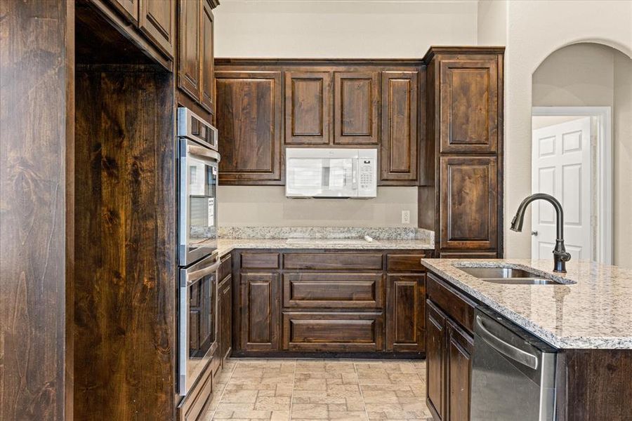 Kitchen featuring stainless steel appliances, dark wood finish cabinets, a kitchen island with sink, light stone finish flooring, and light stone counters