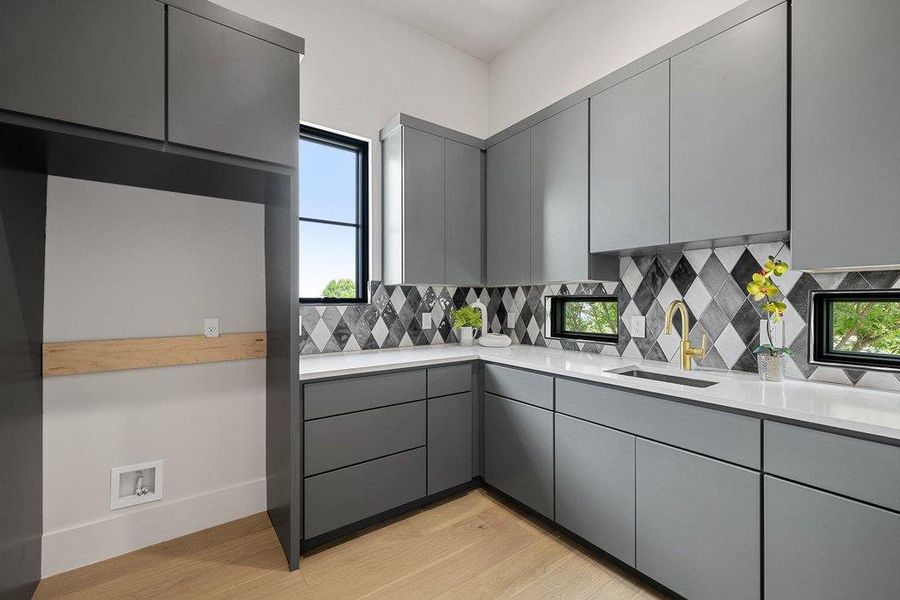Modern utility space featuring light wood-finish flooring, ample grey cabinetry, a striking harlequin tile backsplash, white countertops, and a gold-finish faucet