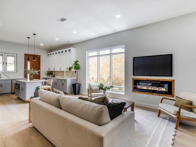 Open-concept living space with light wood-finish flooring, recessed lighting, and a built-in linear fireplace with a wood frame Open-concept living space with light wood-finish flooring, recessed lighting, and a built-in linear fireplace with a wood frame