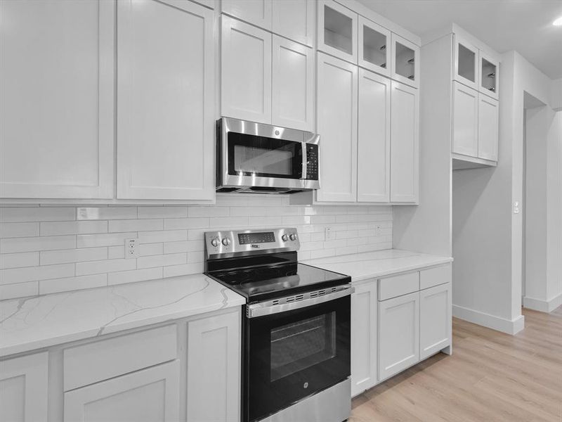 Kitchen featuring stainless steel appliances, tasteful backsplash, light stone countertops, glass insert cabinets, and light wood-type flooring Kitchen featuring stainless steel appliances, tasteful backsplash, light stone countertops, glass insert cabinets, and light wood-type flooring