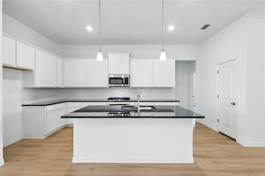 Kitchen featuring an island with sink, dark stone countertops, light wood finished floors, white cabinets, and hanging light fixtures