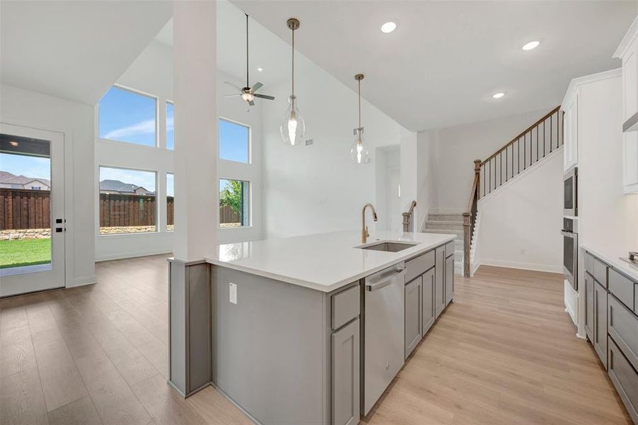 Kitchen island with a white countertop and integrated sink, light wood-finish flooring, white cabinetry, stainless steel appliances, and a staircase with dark wood handrails Kitchen island with a white countertop and integrated sink, light wood-finish flooring, white cabinetry, stainless steel appliances, and a staircase with dark wood handrails