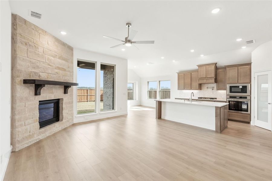 Open living area with stone fireplace, ceiling fan, and kitchen with wood cabinets and center island Open living area with stone fireplace, ceiling fan, and kitchen with wood cabinets and center island