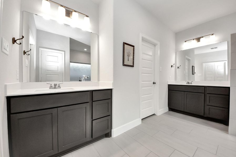 This modern bathroom features dual vanities with dark cabinetry, large mirrors, and bright overhead lighting. Light-colored walls and flooring maintain a clean, minimalistic, and inviting aesthetic.