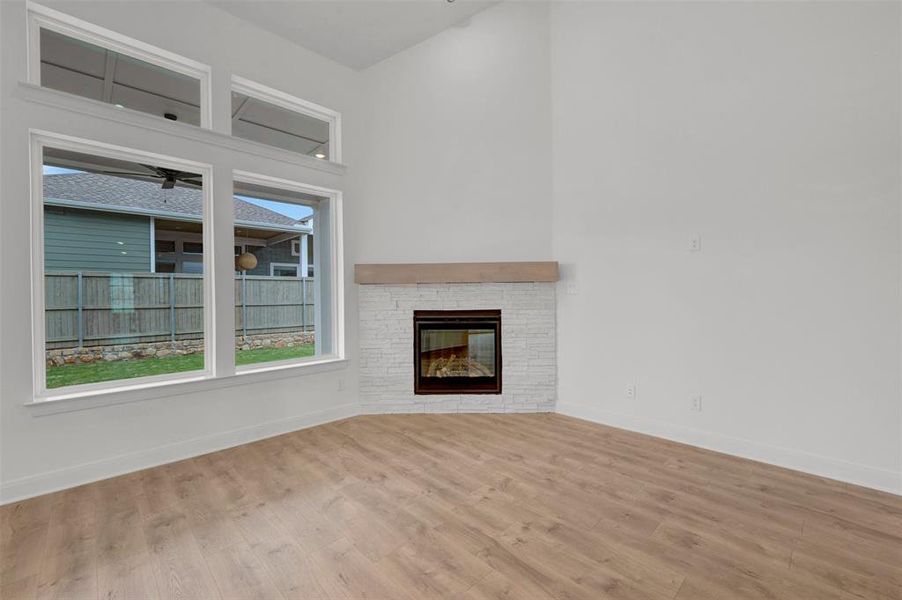 Living area featuring light wood tone flooring, a corner-placed fireplace with a white brick surround and wooden mantel, and large windows providing natural light