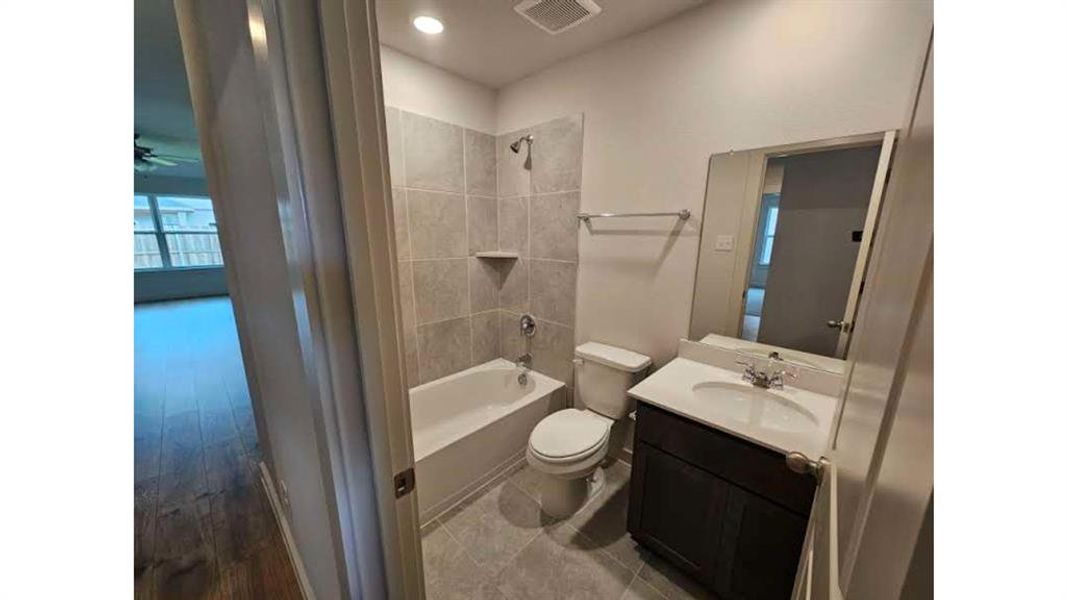 Bathroom featuring a single vanity with a white countertop and dark wood-finish cabinetry, a mirror, a toilet, a tub with a shower, and light gray wall and floor tiling