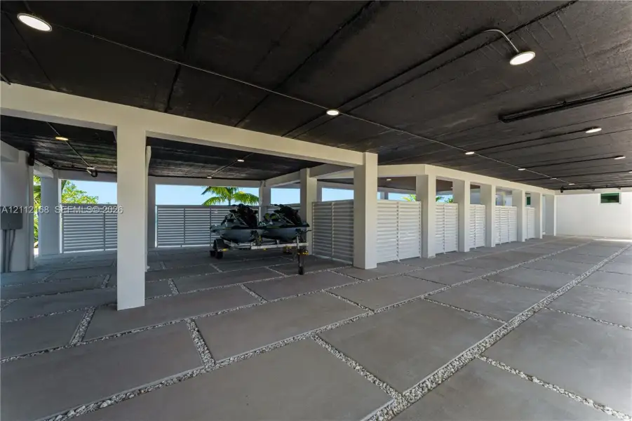 Covered parking area featuring large-format concrete pavers with stone aggregate borders, structural support pillars, white privacy screening, and recessed lighting Covered parking area featuring large-format concrete pavers with stone aggregate borders, structural support pillars, white privacy screening, and recessed lighting