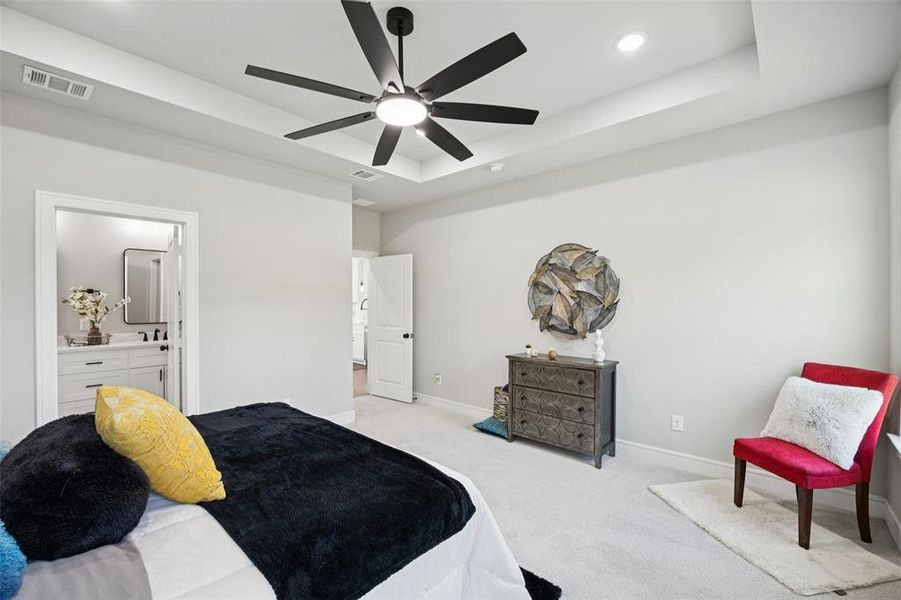 Carpeted bedroom with a raised ceiling, ceiling fan, connected bathroom, and recessed lighting