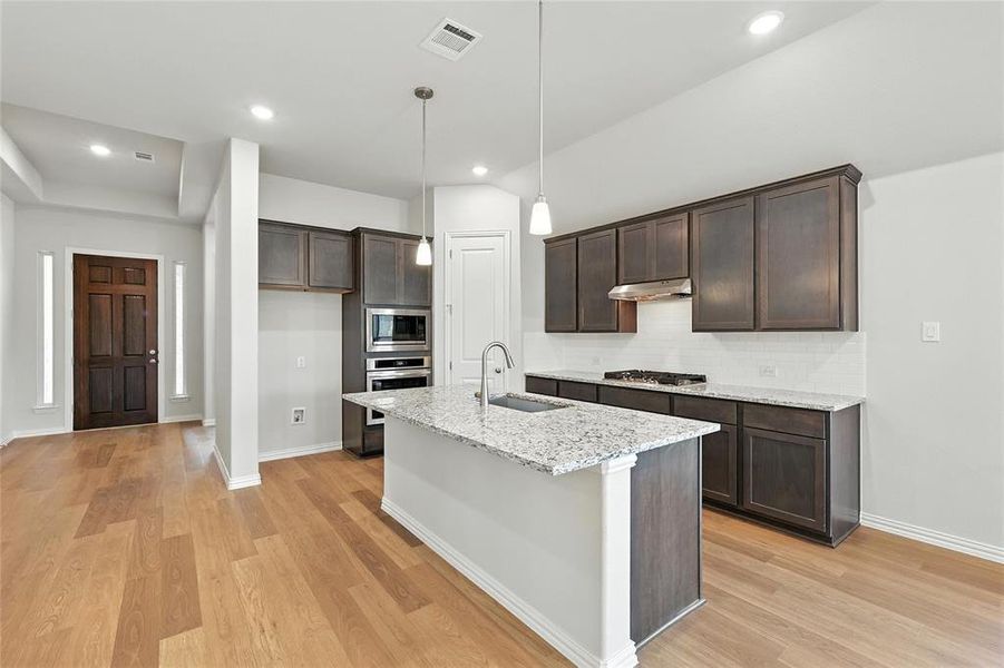 Kitchen with light stone counters, dark wood finish cabinetry, stainless steel appliances, decorative light fixtures, and a center island with sink