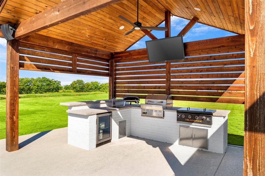 View of patio kitchen with area for grilling, blackstone, sink, ceiling fan, and beverage cooler.