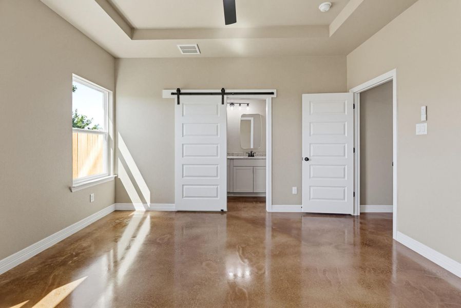 Unfurnished bedroom with a barn door, a tray ceiling, finished concrete floors, ensuite bathroom, and ceiling fan
