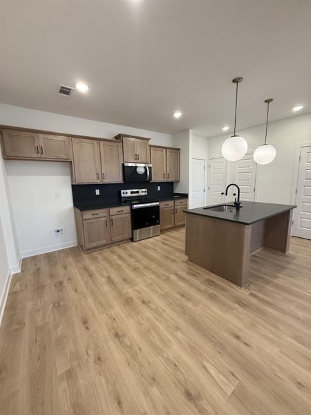 Kitchen featuring dark countertops, stainless steel appliances, an island with sink, decorative light fixtures, and light wood-style floors