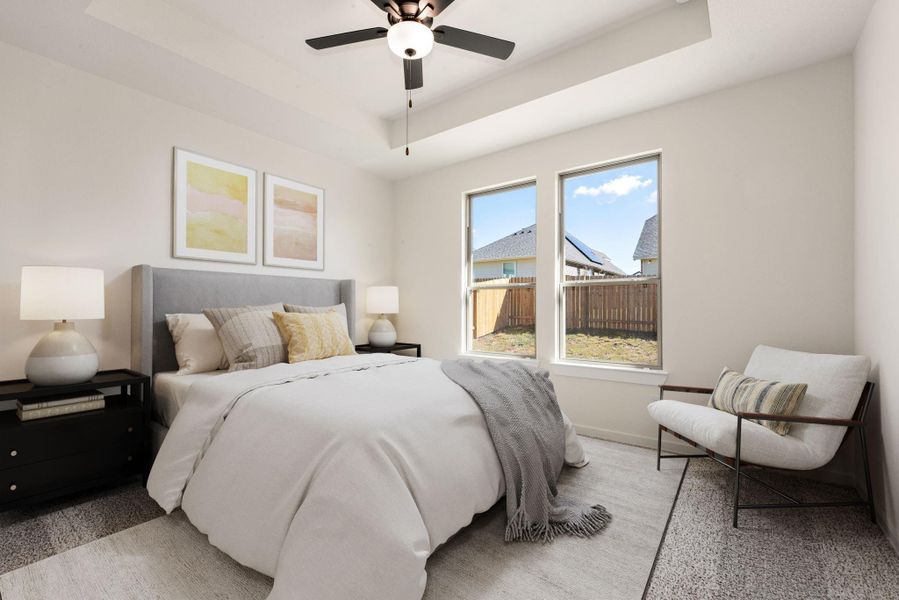 The spacious primary suite is a serene retreat, featuring a striking tray ceiling with a modern ceiling fan and plush carpet flooring throughout. The room is brightly lit by large picture windows that capture the scenic outdoor views. *Virtually Staged The spacious primary suite is a serene retreat, featuring a striking tray ceiling with a modern ceiling fan and plush carpet flooring throughout. The room is brightly lit by large picture windows that capture the scenic outdoor views. *Virtually Staged