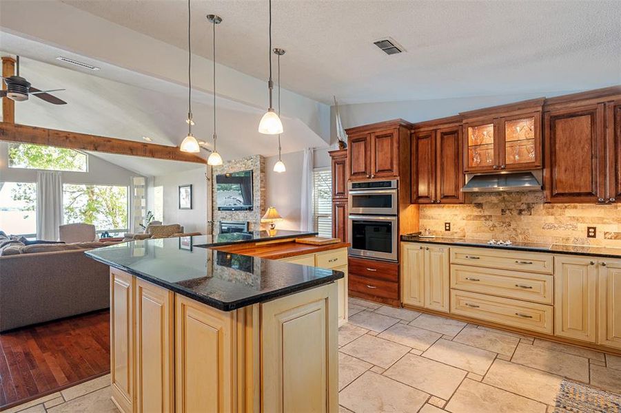 Gourmet kitchen featuring a large island with a black stone countertop, integrated double ovens, warm wood cabinetry, neutral tile flooring, and pendant lighting