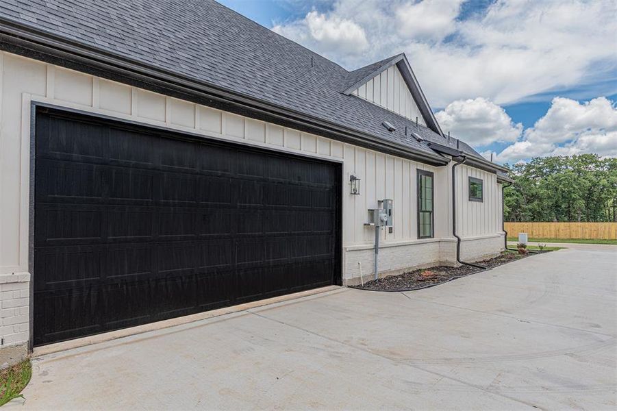 Black panel garage door with a concrete driveway, white board-and-batten siding, dark shingle roofing, and black gutters
