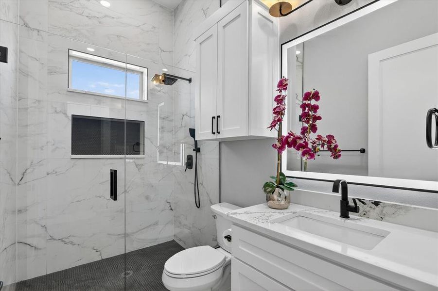Walk-in shower with a glass enclosure, white marble-look wall tiling, and dark floor tiling