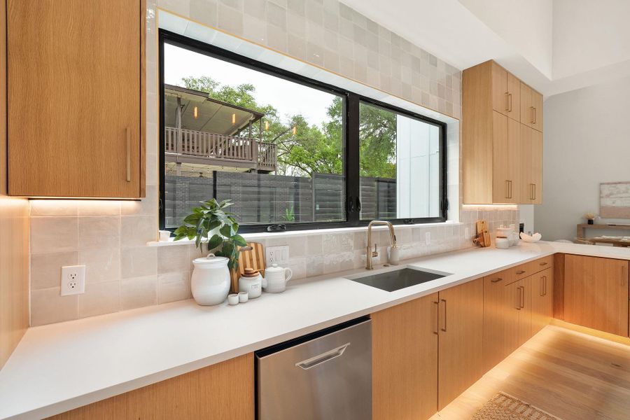 Modern kitchen featuring light wood cabinetry, a large black-framed window, white countertops, square tile backsplash, and stainless steel sink with a gooseneck faucet