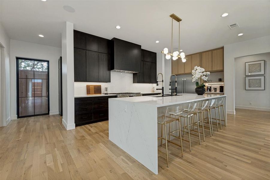 Modern kitchen featuring a waterfall island with a fluted panel base, dark wood cabinetry, integrated appliances, and light wood-finish flooring Modern kitchen featuring a waterfall island with a fluted panel base, dark wood cabinetry, integrated appliances, and light wood-finish flooring