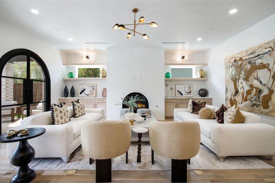 Spacious living area featuring light wood-finish flooring, a contemporary chandelier, recessed lighting, and a white fireplace with a rounded opening Spacious living area featuring light wood-finish flooring, a contemporary chandelier, recessed lighting, and a white fireplace with a rounded opening