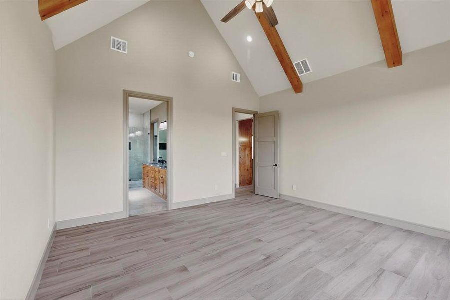 Unfurnished bedroom with high vaulted ceiling, beam ceiling, light wood-type flooring, a ceiling fan, and ensuite bath Unfurnished bedroom with high vaulted ceiling, beam ceiling, light wood-type flooring, a ceiling fan, and ensuite bath