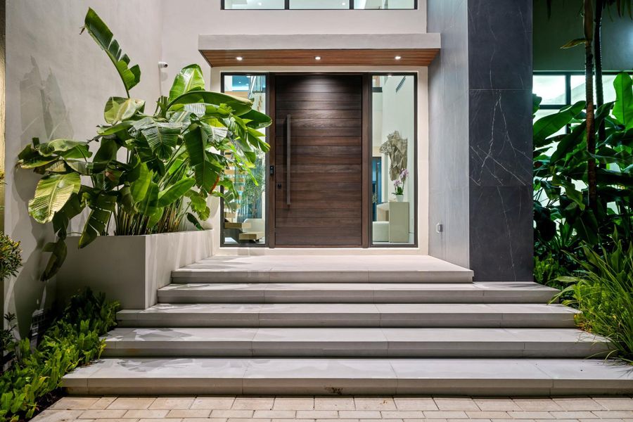 A striking modern entryway with sleek stone steps, tropical landscaping, and a custom wood pivot door framed by floor-to-ceiling glass. A striking modern entryway with sleek stone steps, tropical landscaping, and a custom wood pivot door framed by floor-to-ceiling glass.