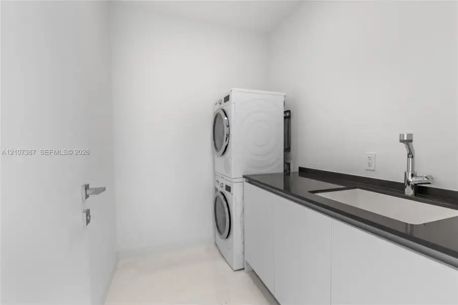 Laundry room featuring stacked washer and dryer, flat-panel cabinetry, dark stone-finish countertop, undermount sink, and high-arc chrome faucet