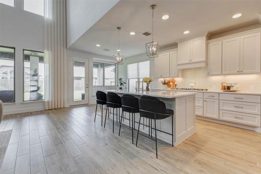 Bright and spacious kitchen with high ceilings, featuring a large island with seating, modern pendant lights, and ample white cabinetry. Large windows and a door open to the backyard, providing plenty of natural light. Bright and spacious kitchen with high ceilings, featuring a large island with seating, modern pendant lights, and ample white cabinetry. Large windows and a door open to the backyard, providing plenty of natural light.