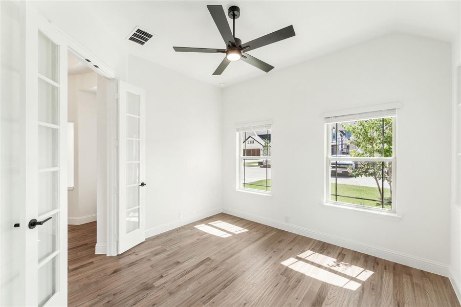 Empty room with hardwood floors, ceiling fan, two windows, and French doors leading to hallway