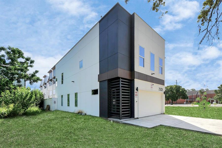 This is a modern two-story building with a clean, geometric design, featuring a mix of flat and protruding facades, large windows, and a garage. It's situated on a lush green lawn with a serene sunset sky in the background.