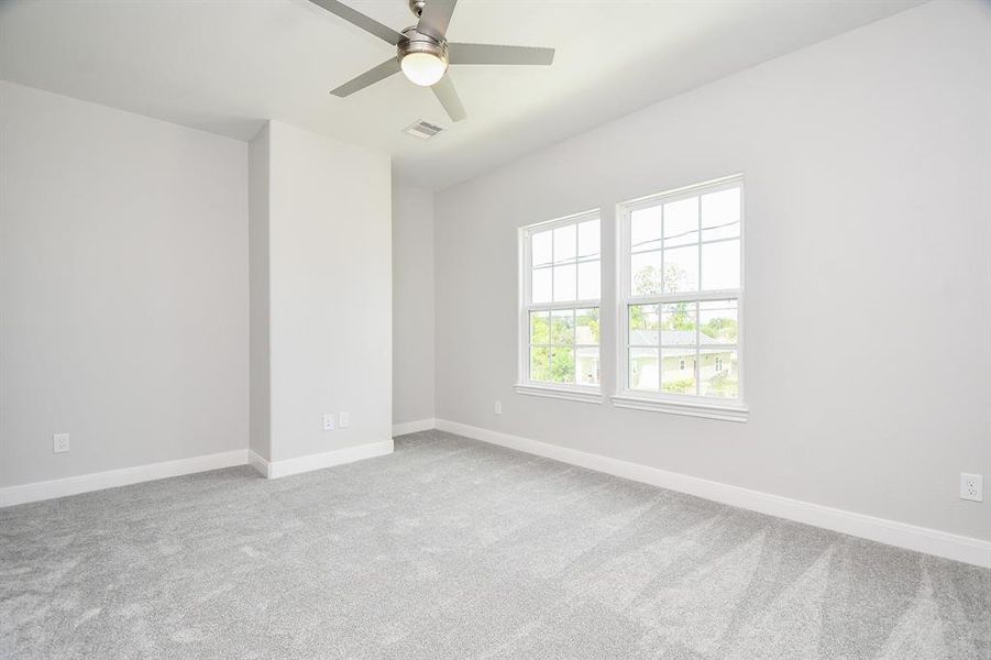 This is a bright, spacious secondary bedroom with a high ceiling, featuring a modern ceiling fan and two large windows providing ample natural light. The room has a fresh look with its neutral walls and new carpet flooring.