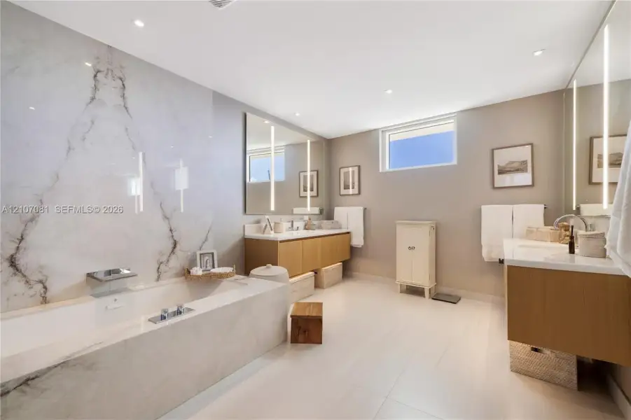 Bathroom featuring a soaking tub with stone-tiled surround, stone slab accent wall, dual floating wood-finish vanities, and vertical integrated mirror lighting Bathroom featuring a soaking tub with stone-tiled surround, stone slab accent wall, dual floating wood-finish vanities, and vertical integrated mirror lighting