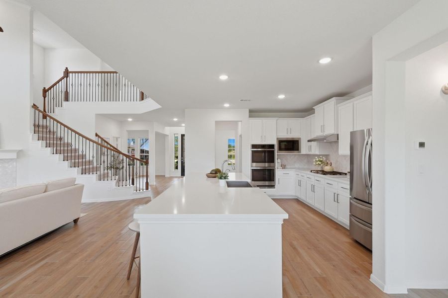 This photo showcases a bright, open-concept kitchen and living area with modern white cabinetry, stainless steel appliances, and a large island. The space features light wood tile flooring and a staircase with elegant railing, adding a touch of sophistication.