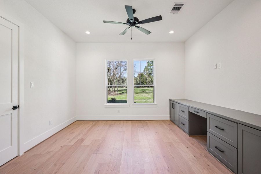 This room features a bright, spacious layout with light wood flooring and a ceiling fan. A large window offers a view of the outdoors, while built-in gray cabinetry provides ample storage space.
