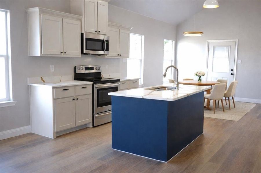 Kitchen featuring stainless steel appliances, an island with sink, light stone counters, vaulted ceiling, and dark wood-style floors Kitchen featuring stainless steel appliances, an island with sink, light stone counters, vaulted ceiling, and dark wood-style floors