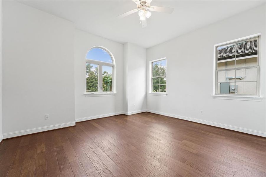 This is a bright, airy bedroom featuring hardwood floors, a high ceiling with a fan, and multiple windows providing natural light, including a distinctive arched window.