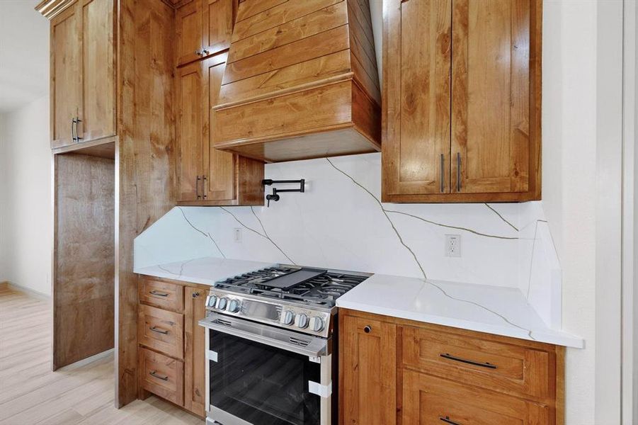 Kitchen featuring stainless steel gas range, brown cabinets, premium range hood, light stone counters, and tasteful backsplash Kitchen featuring stainless steel gas range, brown cabinets, premium range hood, light stone counters, and tasteful backsplash