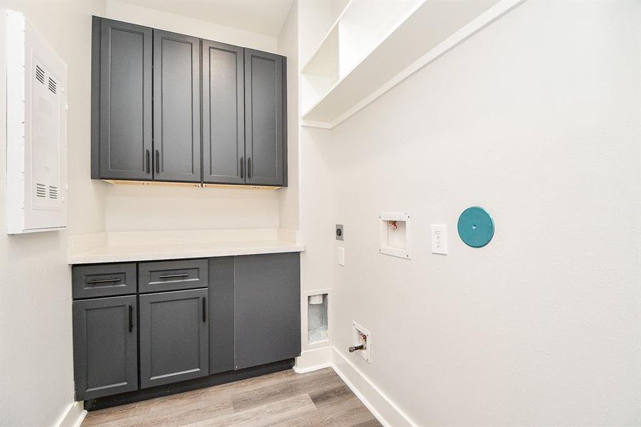 This is a modern utility area featuring dark gray cabinetry, sleek hardware, and light wood countertops, complemented by wood-look flooring and a neutral wall color. There is a space for laundry appliances with necessary hookups, and additional storage options with a wall-mounted cabinet. The room is well-lit and ready for functional use.