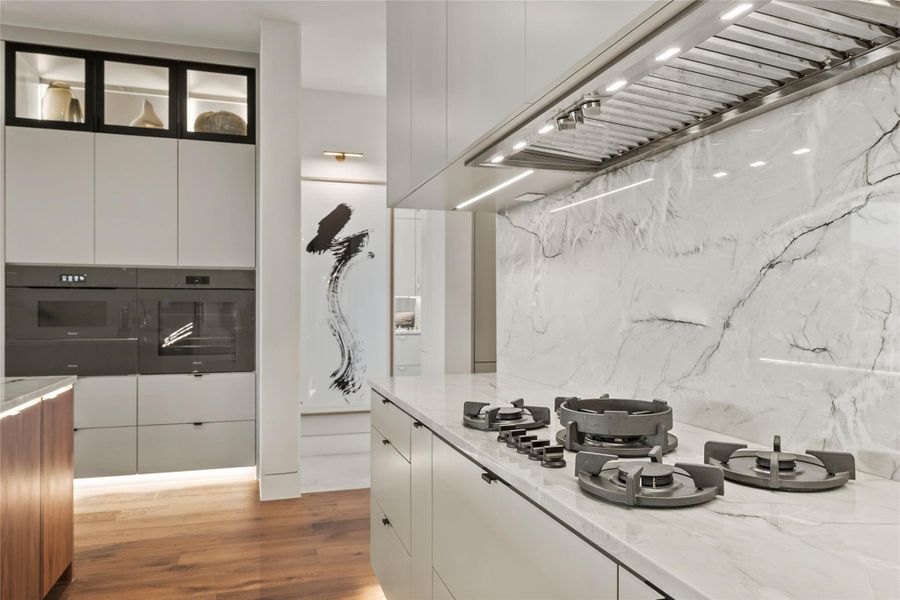 Gourmet kitchen featuring an integrated gas cooktop, sleek range hood, full-height stone slab backsplash, custom cabinetry with display storage, and wood-finish flooring