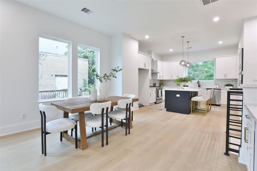 This is a modern, open-plan kitchen and dining area with white cabinetry, stainless steel appliances, and a central island with bar seating. The space is bright with natural light from large windows and features contemporary lighting fixtures.