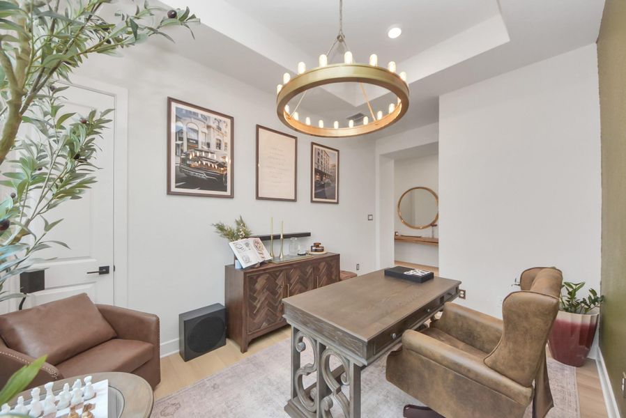Charming home office featuring a stylish chandelier, elegant wooden desk, and cozy leather seating. The space is enhanced by tasteful wall art, abundant natural light, and greenery, creating an inviting and productive atmosphere.