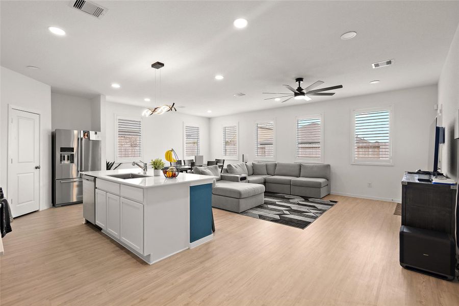 A showcases of a modern, open-concept living area with a spacious kitchen featuring a central island. The living room includes a large sectional sofa and ample natural light from multiple windows. The room is well-lit with recessed lighting and a ceiling fan, complemented by light wood flooring for a cozy and inviting feel.