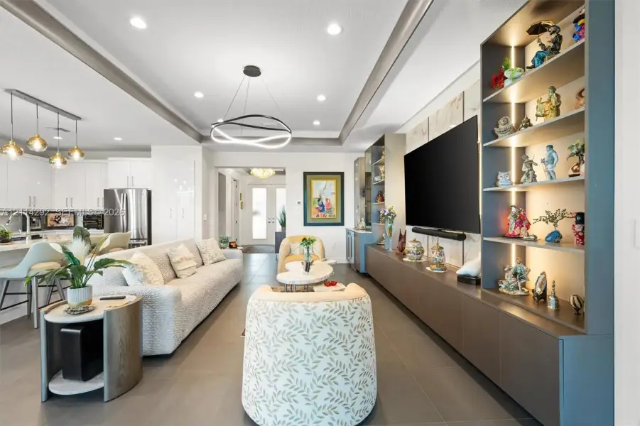 Open-concept living space featuring a recessed tray ceiling with an integrated LED chandelier, large-format tile flooring, and custom built-in cabinetry with illuminated shelving