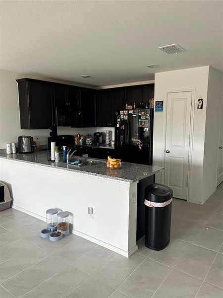 Kitchen with dark stone countertops, fridge, light tile patterned floors, and kitchen peninsula Kitchen with dark stone countertops, fridge, light tile patterned floors, and kitchen peninsula