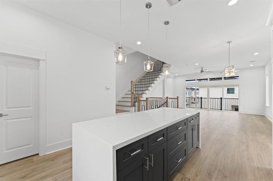 This photo showcases a modern, open-concept living space with a sleek kitchen island, pendant lighting, and light wood flooring. The area features large windows for natural light, a staircase with a contemporary railing, and a spacious living area leading to a balcony.