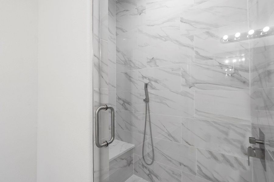 This photo showcases a beautifully upgraded walk-in shower featuring elegant marble-look tile from floor to ceiling. The frameless glass door with brushed nickel hardware adds a sleek, modern touch. Inside, a built-in shower bench provides comfort and convenience, while the handheld showerhead offers versatility.