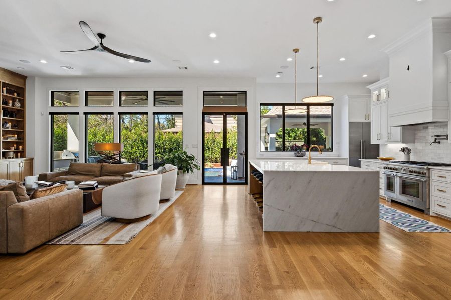 Expansive open-concept living and kitchen with seamless indoor-outdoor flow. Features a statement waterfall-edge island, wide-plank hardwood flooring, recessed lighting, and clean-lined millwork. Walls of windows flood the space with natural light, creating an ideal setting for everyday living and entertaining.