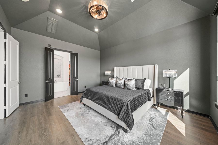 A view of the owner's suite with access to the luxurious private bath through 8-foot double-doors. Warm Revwood flooring and a decorative light fixture with fan add to the elegance of this room.